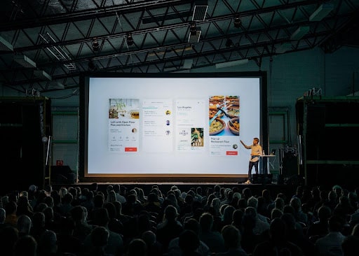 Presenter speaking to an audience with a large screen, representing the need to avoid branded podcast mistakes through effective communication.