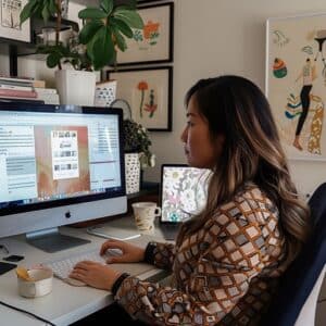 A professional woman engages in B2B podcast planning on her computer in a well-organized home office, surrounded by vibrant art and plants, illustrating the behind-the-scenes effort that goes into creating a business-to-business podcast.