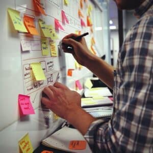 A man in a plaid shirt annotates a whiteboard filled with colorful sticky notes, planning themes and episodes for a B2B podcast, demonstrating the creative and organizational process involved in podcast production.
